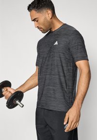 Lyhythihainen harmaa urheilusuoja miehille, jossa on hienovarainen kuvio, pyöreä kaula-aukko ja valkoinen logo; painotettu käsipainoa pidetään oikeassa kädessä.