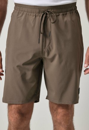Brune elastiske taljeshorts med snøre båret af en person, der står, synlig fra taljen til knæene mod en ensfarvet baggrund.