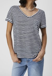 Kvinde iført en navy- og hvidstribet V-hals T-shirt, sølv kædedyse og lyseblå jeans med hænderne i lommerne.