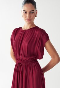 Vestido burdeos con mangas cortas de dolman, cintura ajustada con cordón y textura plisada. Tela suave y fluida en una silueta ligera.