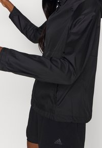 Schwarze leichte Jacke mit Kapuze, glatter Textur, elastischen Bündchen und durchgehendem Reißverschluss. Entworfen für aktive Freizeitbekleidung, kombiniert mit schwarzen Shorts.