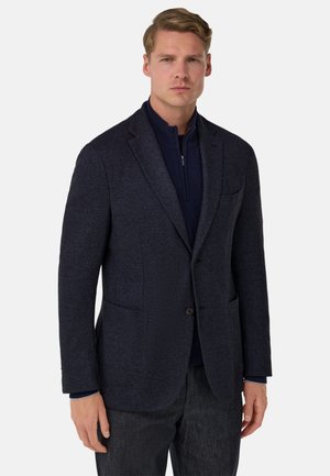 Blazer blu navy a spina di pesce con risvolti a incavo, due tasche frontali e chiusura con un solo bottone, indossato sopra un maglione scuro con zip.