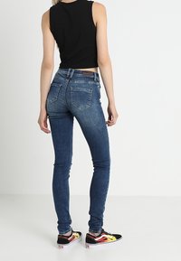 Mujer vestida con vaqueros ajustados azules, top sin mangas negro y zapatillas con patrón de llamas en negro, rojo y amarillo, de pie de espaldas sobre un fondo blanco.