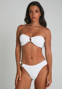 Ensemble de bikini blanc en tissu texturé. Le haut sans bretelles présente un détail en anneau doré au centre ; le bas possède un accent similaire en anneau sur le côté.