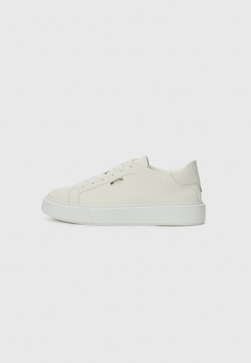 Sneaker bianca a basso profilo con design a lacci, pannello laterale texturizzato e suola spessa, mostrata di profilo su sfondo neutro.