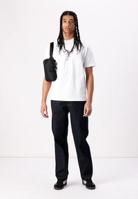 White t-shirt with multi-colored logo, dark blue denim jeans, black shoulder bag, black shoes with white stripes, standing against a white backdrop.