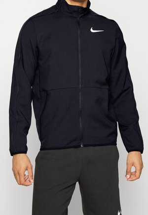 Homme portant une veste zippée noire Nike et un pantalon foncé, debout, les bras détendus le long du corps, devant un fond clair.