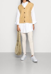 Beżowa dzianinowa kamizelka z dekoltem w serek i pięcioma guzikami, nałożona na białą koszulę zapinaną na guziki. Jasne legginsy i białe trampki.