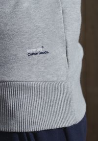 Sudadera gris de algodón con puños acanalados. Presenta un logo azul marino que dice "Superdry Cotton Goods" cerca do do do do.