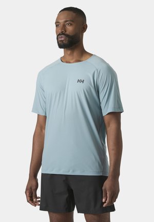 Man draagt lichtblauw atletisch shirt met korte mouwen met "HH"-logo en zwarte korte broek, staand tegen een effen lichte achtergrond.