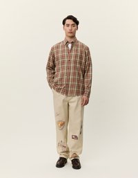 Μακρυμάνικη καρό πουκαμίσα σε καφέ και μπεζ αποχρώσεις, φορεμένη με μπεζ παντελόνι με εκτυπωμένες εικονογραφήσεις τροφίμων. Τα καφέ παπούτσια ολοκληρώνουν το σύνολο.