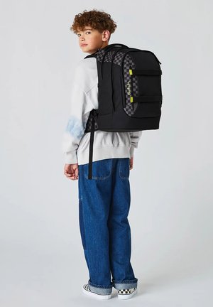 Junge in weißem Sweatshirt und blauer Jeans mit großem schwarzem und kariertem Rucksack, der über die Schulter schaut vor schlichtem Hintergrund.