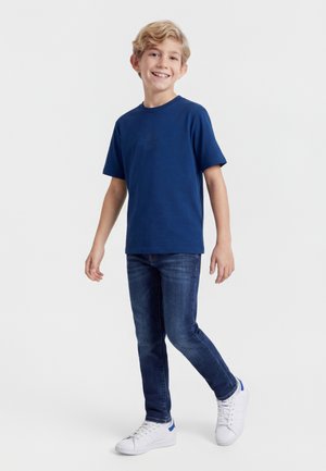 Usmievajúci sa blond chlapec stojaci, oblečený v tmavomodrom tričku, tmavomodrých džínsoch a bielych teniskách s modrými detailmi na jednoduchom pozadí.