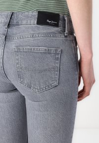 Grå denim jeans med en tettsittende design, en baklomme med et subtilt søm-mønster, og en svart etikett ved livlinjen.