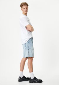 Hvid t-shirt med korte ærmer, lyseblå denim shorts og sorte sko. Modellen står sidelæns med armene krydset og viser outfittet.