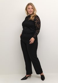 Svart långärmad topp med spetsmönster, kombinerad med svarta smalbyxor. Modellen bär svarta spetsiga klackskor, totalt en mjuk yta.