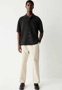 Camisa de botones negra de manga corta con un bolsillo, combinada con pantalones ligeros beige y zapatos negros. Diseño simple y limpio con un ajuste relajado.