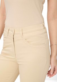 Pantalons moulants beiges en tissu doux, avec des poches en biais, une fermeture éclair avec un bouton et des détails cousus le long de la taille.