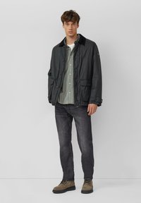 Zwarte, lichtgewicht jas met een corduroy kraag, twee voorzakken, gedragen over een groen geruit overhemd, gecombineerd met donkere jeans en bruine laarzen.