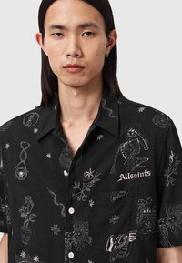 Μαύρο κοντομάνικο πουκάμισο με λευκή περίτεχνη κεντητική που παρουσιάζει διάφορα σχέδια, συμπεριλαμβανομένων κρανίων και αστεριών, πάνω σε υφή υφάσματος.