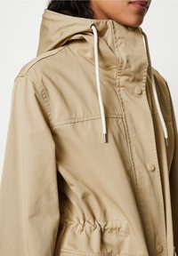 Kvinna i beiget huva-jacka med vita dragsnören och tryckknappar, närbild av övre torso och nacke.