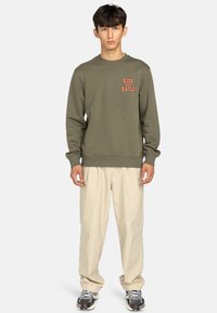 Sudadera verde oliva con puños y dobladillo acanalados, con la frase "RIDE THE RAILS" impresa en naranja. Combinada con pantalones holgados beige y zapatillas.