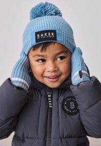 Svetlo modra pletena kapa s cofkom in značko z logotipom; ujemajoče modre črtaste rokavice; siva puhovka z okroglo značko z logotipom.