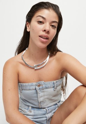 Collar de metal plateado con un diseño curvado y abierto que presenta dos acentos centrales. Se lleva con un top de tubo de mezclilla. La piel es visible en los hombros.