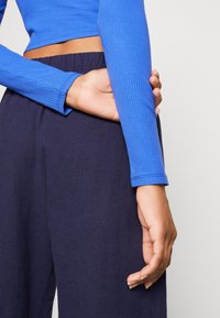 Långärmad, ruggad topp i ljusblå, kombinerad med mörkblå, löst sittande byxor. Handen vilar på midjeområdet.