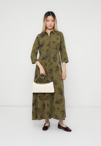 Abito lungo verde oliva con stampa floreale scura, chiusura frontale con bottoni, maniche a tre quarti e vita arricciata. Accompagnato da una borsa bianca.