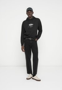 Μαύρο φούτερ με κουκούλα και γραφικό τύπωμα, μαύρο παντελόνι με άνετη εφαρμογή και λευκά αθλητικά παπούτσια. Αξεσουάρ περιλαμβάνουν σκούφο σε σκούρο χρώμα και μαύρη ζώνη.