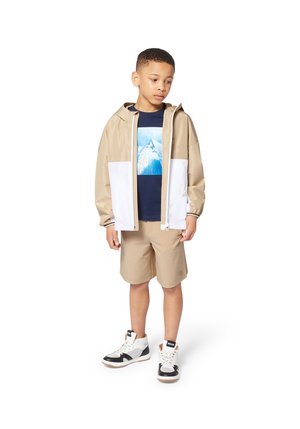 Beige en witte windbreaker met capuchon, gecombineerd met een marineblauwe graphic t-shirt en beige shorts. Schoeisel: hoge sneakers in zwart en wit.