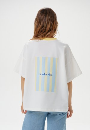 Donna con i capelli lunghi che indossa una maglietta bianca oversize con strisce blu e gialle e il testo "la dolce vita" sulla schiena, abbinata a jeans azzurri.
