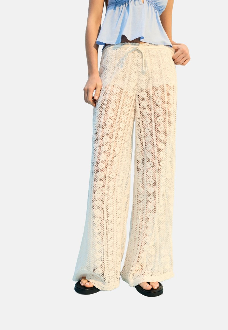 Pantalons larges en crochet blanc avec des motifs verticaux et une taille à cordon. Associés à des sandales noires. Tissu doux et texturé.