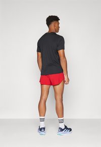 Schwarzes Sport-T-Shirt, rote Shorts, weiße Socken mit schwarzen Streifen und blaue Laufschuhe mit hellgrünen Akzenten. Das Modell wird von hinten gesehen.