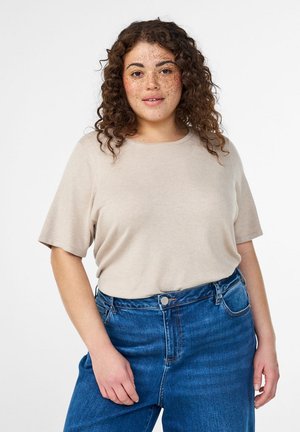 Lockige Frau mit Sommersprossen trägt ein beiges kurzärmeliges Oberteil und blaue hoch taillierte Jeans, steht mit einer Hand nahe ihrer Tasche.