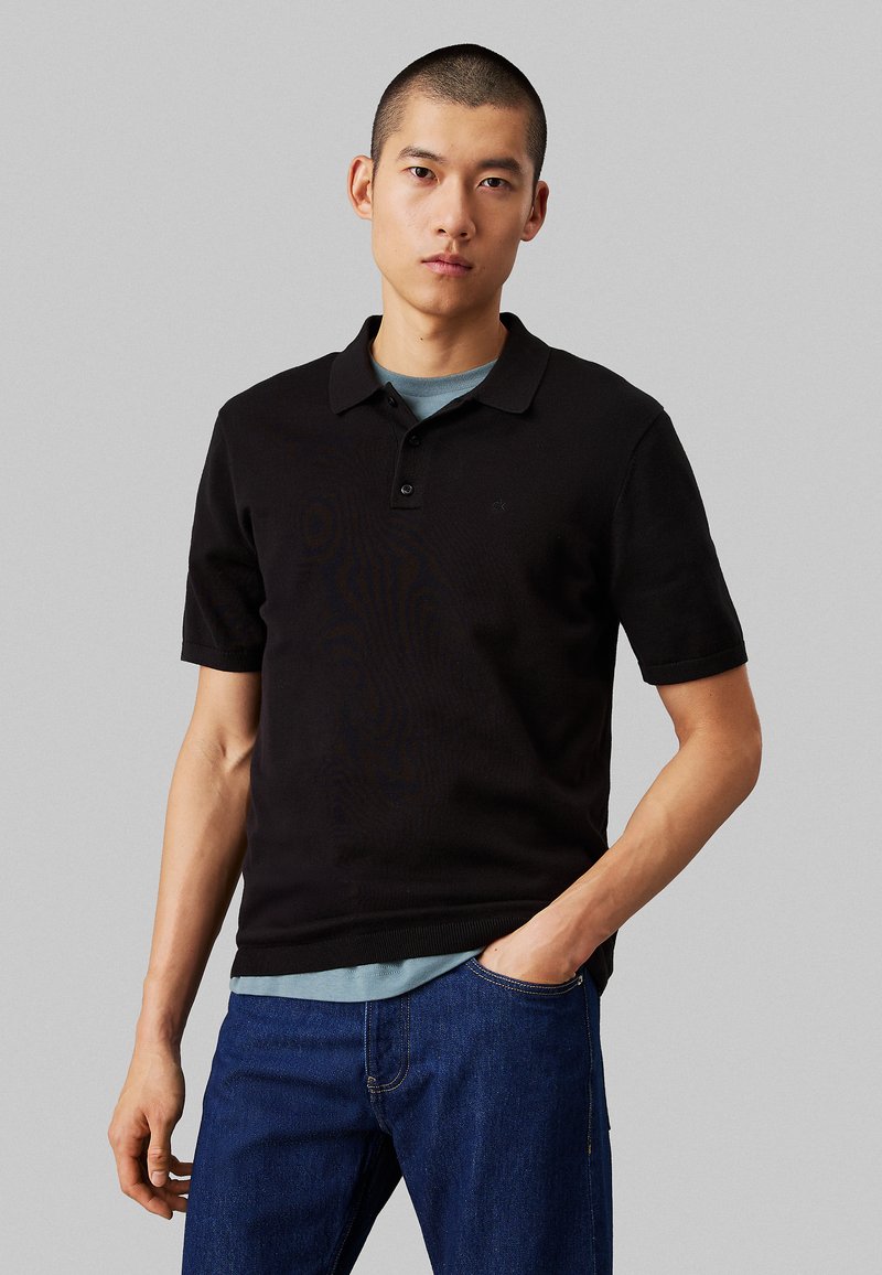 Polo shirt nero in cotone, con colletto classico, maniche corte e patta con due bottoni. Indossato sopra una maglietta azzurra chiara.