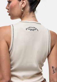 Top senza maniche color beige chiaro con una texture a coste, dettagli di cucitura nera e un logo "Good For Nothing" ricamato sul retro.