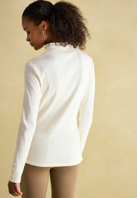 Valge ribakujuline pika varrukaga topp, millel on volangkaelus ja nööpdetailid varrukatel. Sile kangas, mis rõhutab figuuri.