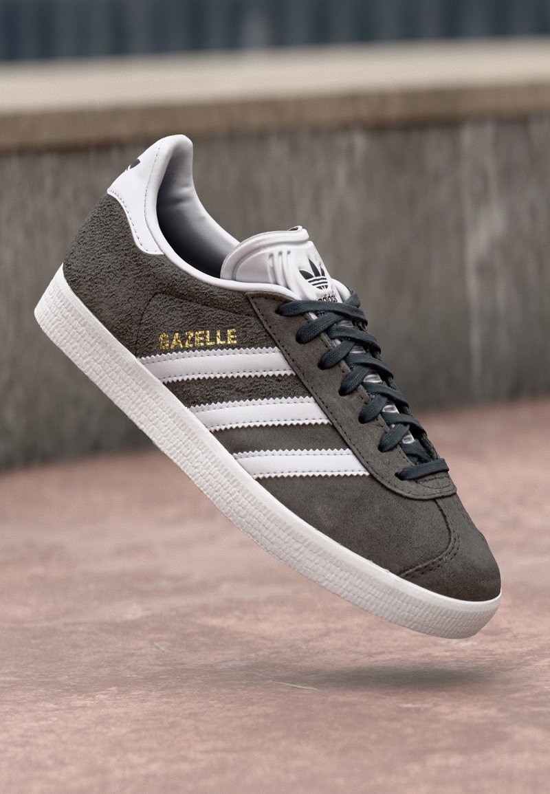 Grå mockasinsneaker med vita läderränder och guldfärgad "Gazelle"-text. Har svarta skosnören och texturerad vit gummisula.