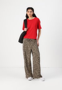 Top rosso a maniche corte abbinato a pantaloni larghi beige con stampa leopardata e sneakers bianche. La modella tiene una borsa nera su una spalla.