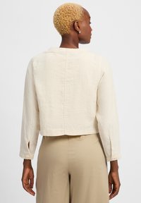 Cropped beige jacket made of lightweight fabric, featuring long sleeves, a straight hem, and buttoned cuffs. Smooth texture, minimal design.