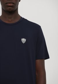 Tamsiai mėlynas medvilninis marškinėliai su apvaliu kaklu, trumpomis rankovėmis ir mažuoju siuvinėtu EA7 logotipu šarvo dizaine kairėje krūtinės pusėje.