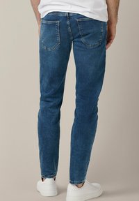 Niebieskie dżinsy o dopasowanym kroju, z pośrednim stanem, dwiema tylnymi kieszeniami i czystym wykończeniem. Noszone z białymi trampkami.