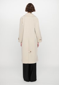 Beige long coat with a collar, side pockets, and large buttons. Features a clean, minimalist design and a slightly textured fabric.