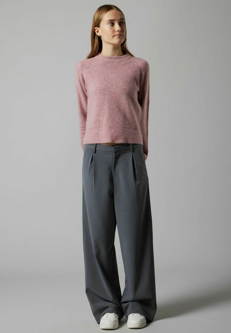 Un pull tricoté rose clair à manches longues, associé à un pantalon large gris sur mesure et des baskets blanches, se tenant devant un fond neutre.