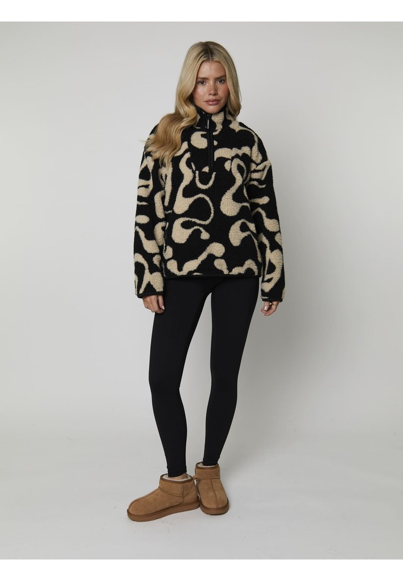 Pull en polaire noir et crème avec un motif abstrait, col haut et fermeture éclair sur le devant. Associé à des leggings noirs et des bottes beiges.