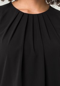 Sort plisseret bluse med bådhals, blød tekstur og subtil drapering, der fremhæver det slanke, minimalistiske design.