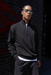 Black zip-up sweatshirt with a high collar, featuring a silver zipper and a blue accent. Worn over a white t-shirt, paired with dark cargo pants.