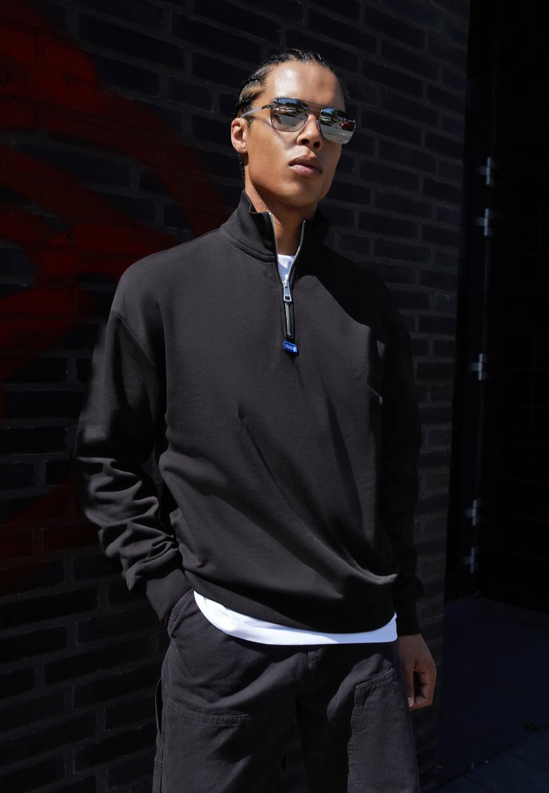 Black zip-up sweatshirt with a high collar, featuring a silver zipper and a blue accent. Worn over a white t-shirt, paired with dark cargo pants.